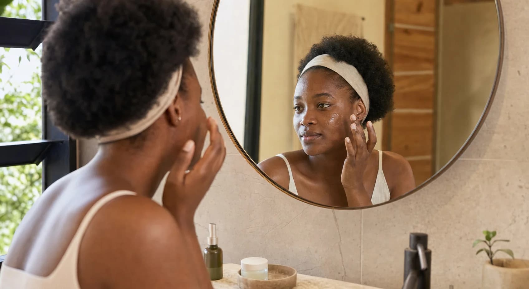 A Nigerian woman applying lightweight skincare while looking in a bathroom mirror